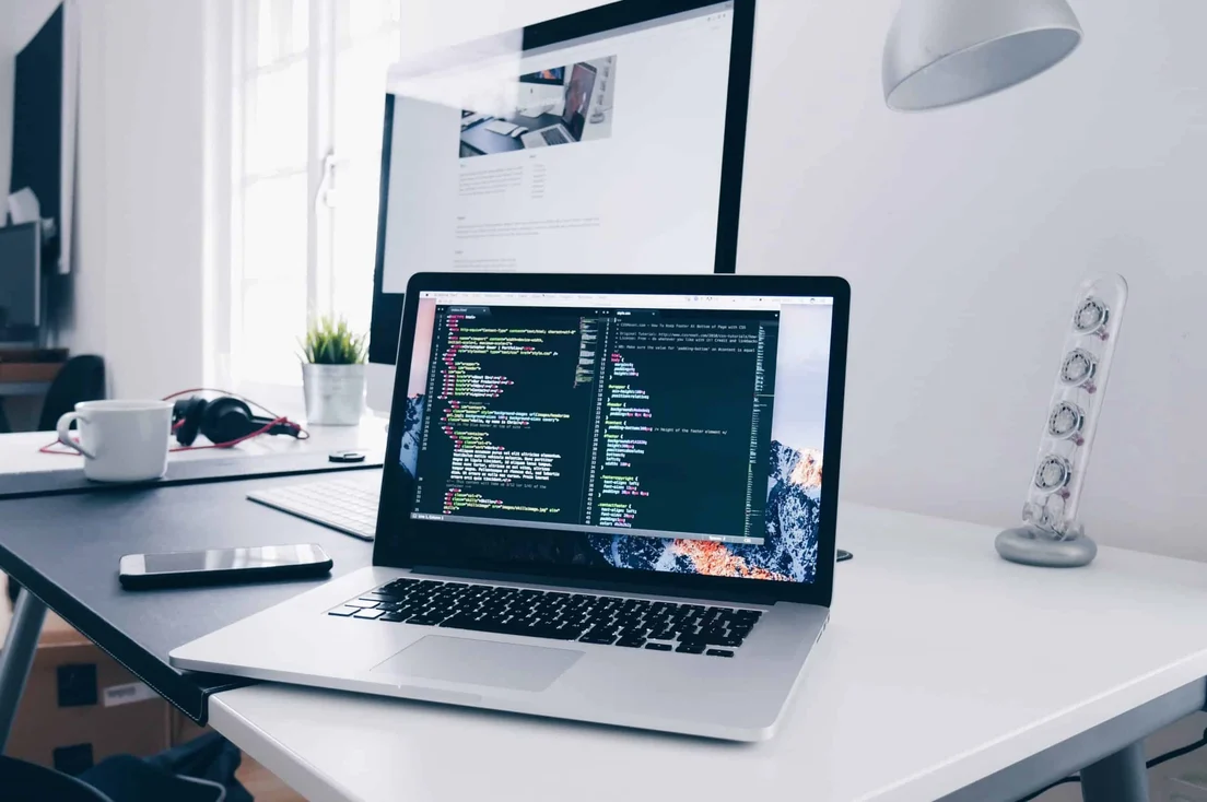 A laptop showing code on a desk beside a monitor.