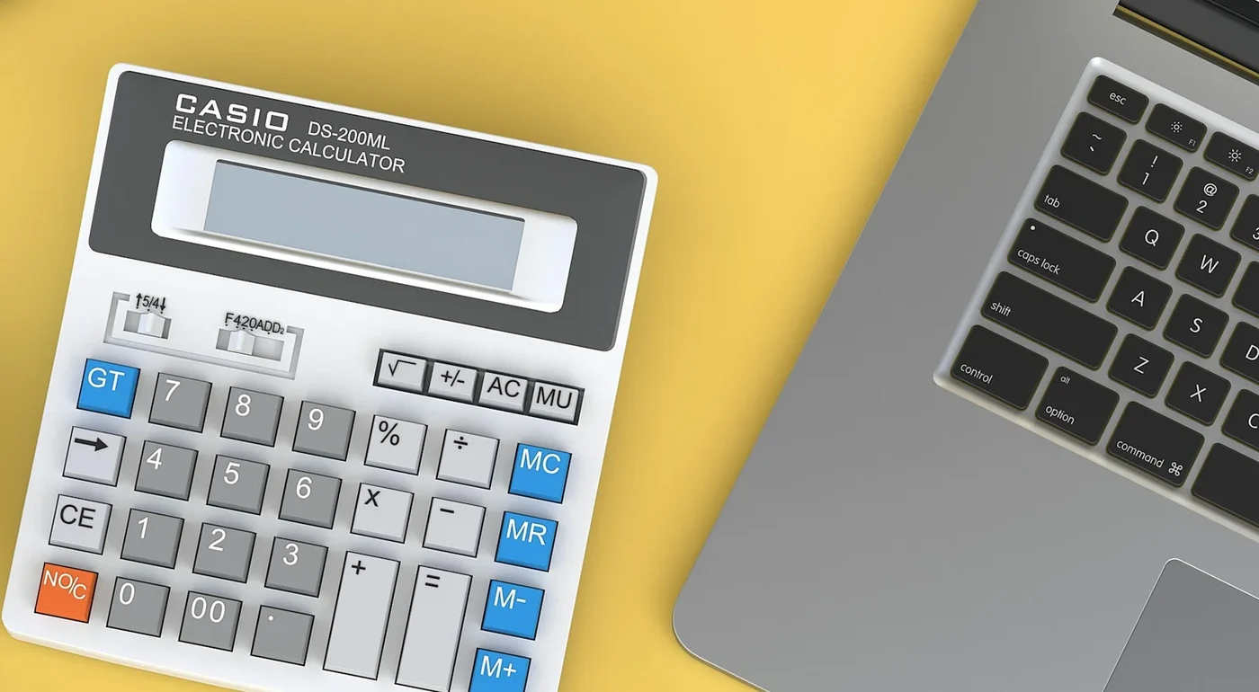 A desk calculator beside an open laptop on a yellow desk.